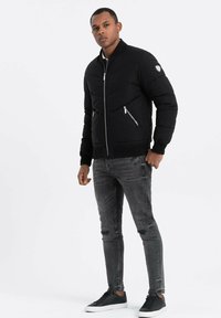 Svart vadderad bomberjacka med hög krage, dragkedja fram, två sidofickor med dragkedja och ribbade manschetter, tillsammans med grå slitna jeans och svarta sneakers.