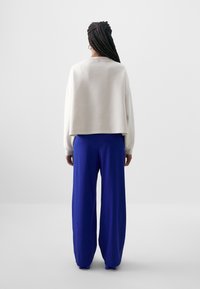 Sweatshirt cropped cinza claro com detalhe de botão nas costas, combinado com calças largas azuis brilhantes, apresentado de uma vista traseira.