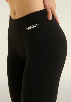 Gros plan sur une personne portant des leggings noirs Freddy, avec une main reposant sur la hanche, montrant le logo Freddy sur la ceinture.