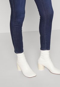 Jeans ajustados de denim azul oscuro combinados con botas tobilleras de imitación de piel blanca con un diseño arrugado y tacón grueso.