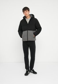 Pufferjakke i sort og grå, med lynlås foran, hætte og polstret design. Bæres over en let sweater og sammen med sorte jeans.