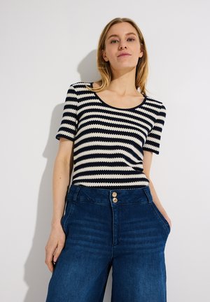 Frau trägt ein schwarz-weiß gestreiftes Kurzarmoberteil mit horizontalen Streifen, in dunkelblaue hochtaillierte Jeans gesteckt und steht mit einer Hand in der Tasche.