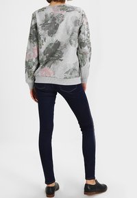 Kobieta w szarej bluzie z zielonymi i różowymi wzorami liści, w ciemnych obcisłych dżinsach i czarnych sznurowanych butach, stojąca tyłem.