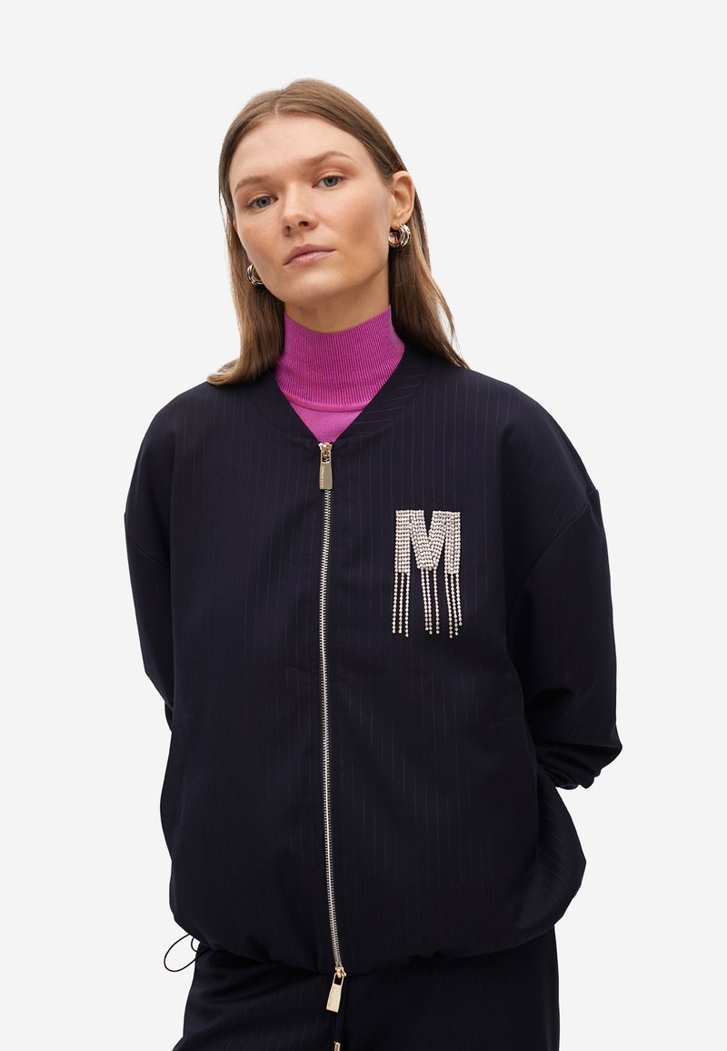 Veste de bombardier navy avec motif à rayures, fermeture éclair argentée et accent "M" orné de cristaux. Portée sur un col roulé magenta.