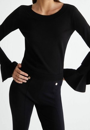 Femme portant un haut noir ajusté avec des poignets larges évasés et un pantalon noir taille haute avec des détails boutonnés.