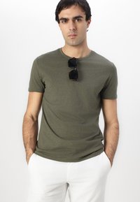 Olivgrünes Kurzarm-T-Shirt aus weichem Stoff, mit rundem Ausschnitt und Slim-Fit. Schwarze Sonnenbrille liegt auf dem Shirt.