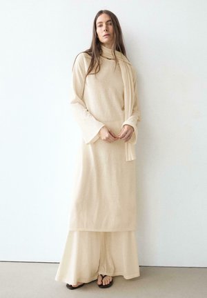 Frau mit langen Haaren, die ein beiges langärmeliges Kleid, einen passenden Schal, weite Hosen und schwarze Sandalen trägt, steht vor einer weißen Wand.