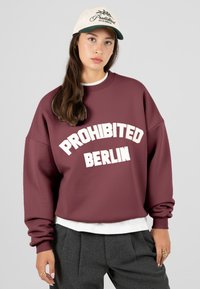 Mladá žena v vínovém svetru s nápisem "PROHIBITED BERLIN", šedých kalhotách, bílém klobouku a zlatých prstenech.