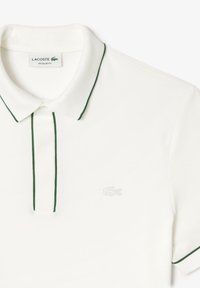 Vit polo t-shirt med gröna detaljer på kragen och knappslån. Har Lacoste-logotypen broderad i ljusgrått på bröstet. Normal passform.