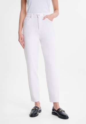 Femme portant un jean blanc taille haute et des mocassins en cuir noir avec détail métallique, debout avec une main dans la poche.