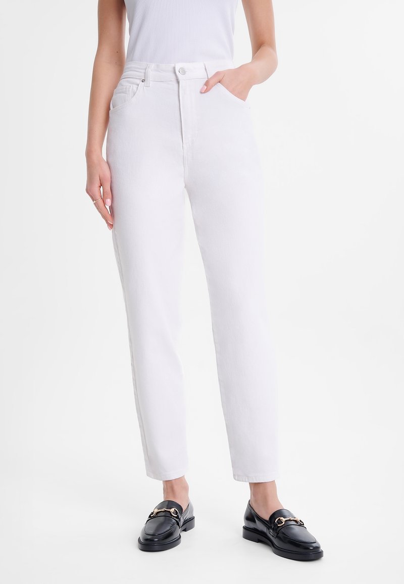 Femme portant un jean blanc taille haute et des mocassins en cuir noir avec détail métallique, debout avec une main dans la poche.