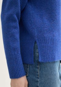 Blauwe trui met geribde manchetten en zoom; heeft een gestructureerde afwerking en zijsplitten. Gecombineerd met denim jeans.