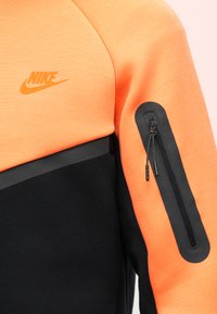 Πορτοκαλί και μαύρο φούτερ Nike με τσέπη με φερμουάρ στον επάνω μανίκι και το λογότυπο της Nike τυπωμένο στο στήθος.