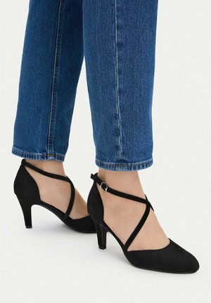Woman wearing black suede pointed-toe heels with crisscross straps paired with blue cropped denim jeans on a white background.
