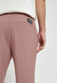 Světle růžové kalhoty zobrazené zezadu, s dvojitými zadními kapsami a černou etiketou. Tkanina vypadá hladce a elegantně.