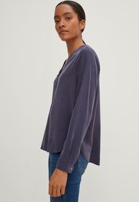 Blusa a maniche lunghe di un profondo viola con scollatura a V e vestibilità rilassata. Tessuto morbido con una texture liscia e polsini con bottoni. Indossata con jeans blu.
