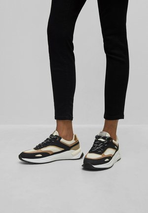 Personne portant des baskets beige, noires et blanches avec des lacets noirs et un pantalon noir ajusté, sur un fond gris uni.