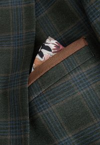 Dark green plaid blazer pocket with a brown trim holding a multicolored floral silk pocket square.