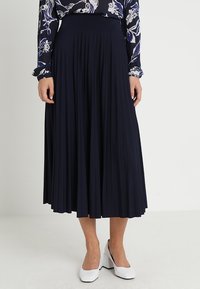 Gonna midi plissettata blu navy con vita alta, realizzata in tessuto morbido, abbinata a scarpe con tacco blocco bianche. Blusa stampata visibile nella parte superiore.