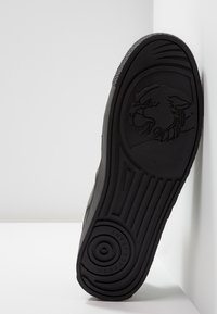 Semelle de chaussure en caoutchouc noir avec tête de Méduse embossée et marque Versace Jeans, inclinée contre un fond blanc en coin.