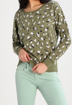 Vrouw draagt een olijfgroen shirt met lange mouwen en luipaardprint en lichtgroene mintkleurige broek, terwijl ze de zoom van het shirt met haar handen recht trekt.