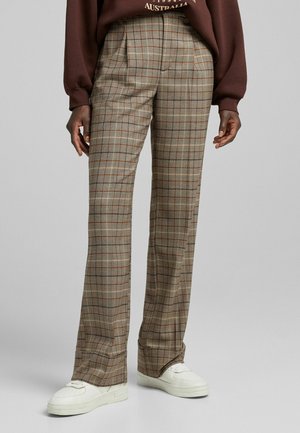 Personne portant un pantalon à carreaux taille haute, jambes larges, tons beige et marron, avec des baskets blanches, debout devant un fond uni.