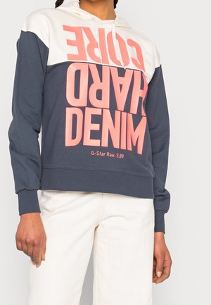 Osoba ubrana w kolorową bluzę z kapturem z różowym napisem „HARD CORE DENIM” i białe spodnie, stojąca na jednolitym tle.