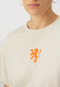 Bež majica iz bombaža s oranžnim vezanim logotipom leva na levem prsnem delu, okroglo ovratnico in kratkimi rokavi.