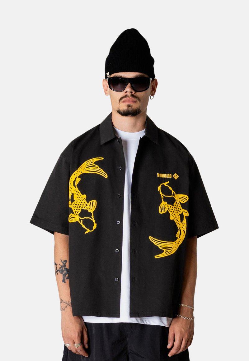 Homme portant une chemise noire à manches courtes avec des motifs de poissons koï jaunes, un débardeur blanc, un bonnet noir, des lunettes de soleil et des tatouages visibles sur le bras.