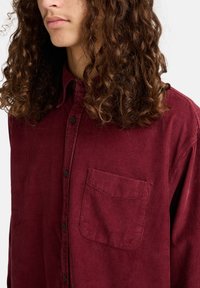 Bordeauxfarbendes Kordhemd mit Button-Down-Kragen, Fronttasche und strukturierten Rillen. Mit schwarzen Knöpfen und einem lässigen Schnitt.