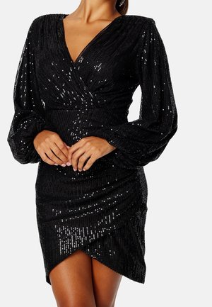 Robe noire à manches longues en sequins avec col en V, taille froncée, manches bouffantes et ourlet asymétrique portée par une personne les mains jointes à la taille.