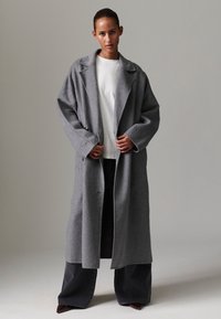 Ilgas pilkas vilnonis paltas su įpjauta apykakle, trimis mygtukais ir platėjančiomis rankovėmis. Derinamas su balta palaidine ir tamsiomis plačiomis kelnėmis.