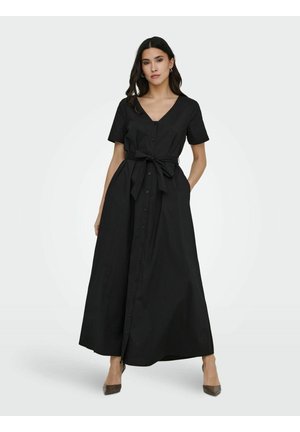 Femme portant une robe maxi noire à manches courtes avec boutons, ceinture nouée à la taille et talons à bout pointu, debout avec une main dans la poche.