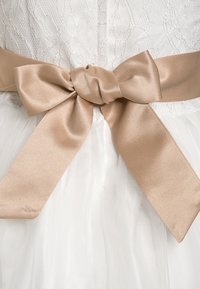 Bílá krajková šaty s průsvitnou tylovou vrstvou, stažená v pase měkkou saténovou béžovou mašlí. Viditelné složité krajkové vzory.