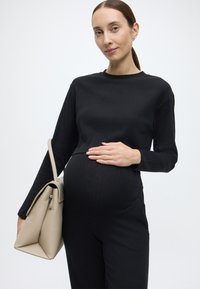 Top de manga comprida preto com nervuras e calças combinando, usados por uma pessoa que exibe uma barriga redonda e grávida. Mala de mão bege claro com design angular.