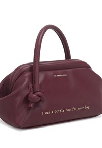 Borsa a mano in pelle vegana bordeaux con manico annodato, con testo metallico che recita: "Ero una bottiglia, ora sono la tua borsa." Texture liscia, forma compatta.