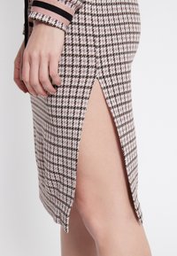 Jupe à motif pied-de-poule en rose, marron et blanc avec une fente sur le côté, fabriquée dans un tissu en tricot texturé et dotée de poignets rayés contrastants.