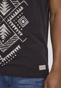 Black t-shirt featuring a beige geometric pattern, including triangles and diamonds. Tag on the bottom left reads "ISOLID Denmark."
