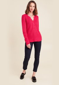 Roten Strickpullover mit V-Ausschnitt und Knopfleiste, der über strukturierte Muster verfügt. Kombiniert mit dunklen engen Jeans und schwarzen Loafers.
