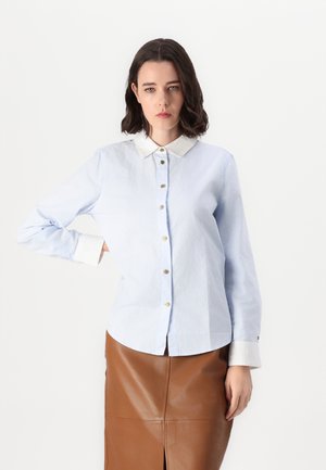 Chemise bleu clair avec un col blanc, dotée de fermetures à boutons dorés et de manches longues. Tissu lisse avec des poignets de manches contrastants.