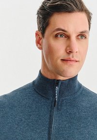 Granatowy sweter zapinany na zamek z wysokim kołnierzem, prążkowaną fakturą i kontrastującym uchwytem do zamka. Gładki materiał, dopasowany krój, minimalistyczny styl.