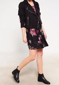 Veste de motard courte en daim noir avec quincaillerie argentée, portée sur une robe noire à fleurs avec des fleurs rouges et grises. Bottines noires.