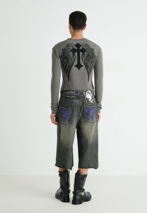Maglietta grigia a maniche lunghe con grafica nera di ali e croce; pantaloni corti di jeans scoloriti con ricami viola; stivaletti neri alla caviglia con fibbie.