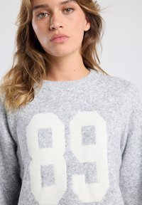 Jeune femme aux cheveux blonds ondulés portant un pull gris clair avec de grands chiffres blancs "89" sur le devant, fond neutre.