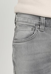 Grå denimjeans med en slät yta, som har en framficka, metallbeslag och sydda detaljer längs midjan och sömmarna.