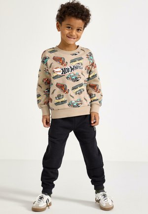 Béžová mikina s barevnou grafikou aut a nápisem „Hot Wheels“; doplněná o černé tepláky jogger a bílé tenisky s hnědými detaily.