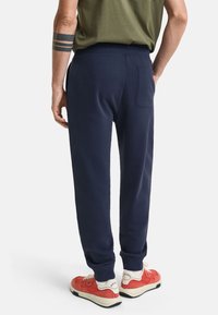 Marineblå joggebukser med myk tekstur, smal passform, elastisk midje og baklomme. Brukt med røde og hvite sneakers.
