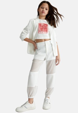 Blazer bianco sopra una t-shirt grafica corta. Pantaloni jogger bianchi con pannelli in rete trasparente. Sneakers bianche con accenti testurizzati.