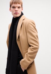 Beige wool overcoat with notched lapels and two front pockets, layered over a black turtleneck and a ribbed black scarf.