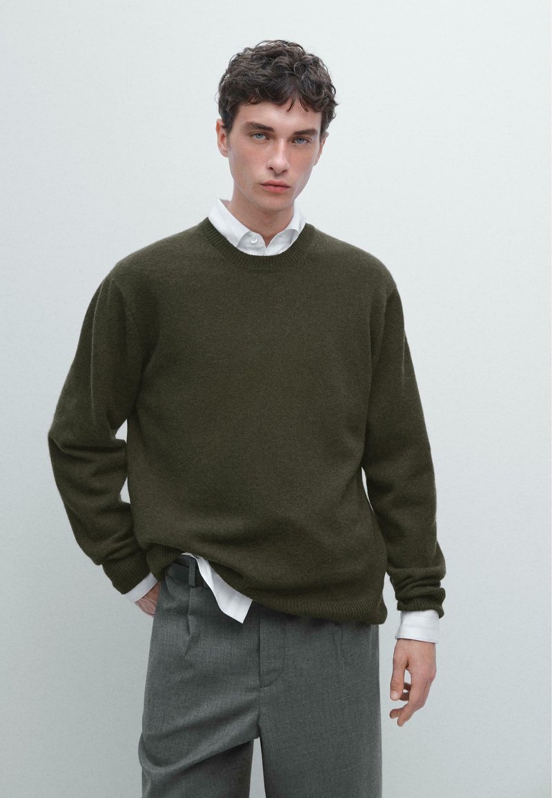 Tmavě olivově zelený pletený svetr s kulatým výstřihem, dlouhými rukávy a volným střihem, doplněný bílou košilí s límečkem a šedými kalhotami.